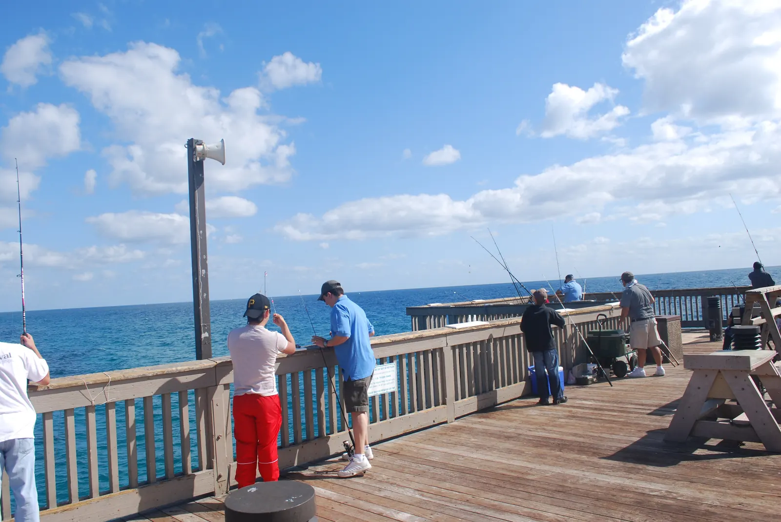 Anglers lean on the rail of the Deerfield Beach pier with rods over the side, blue ocean behind them and condominium towers on the horizon.