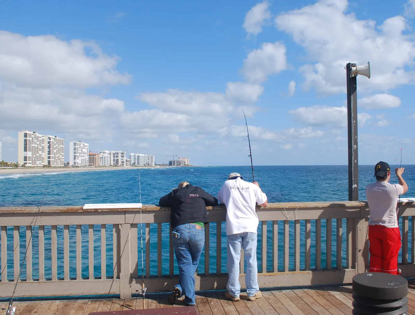 Three people stand at the wooden rail of the Deerfield Beach pier watching their lines, with the coastline and towers to the south.