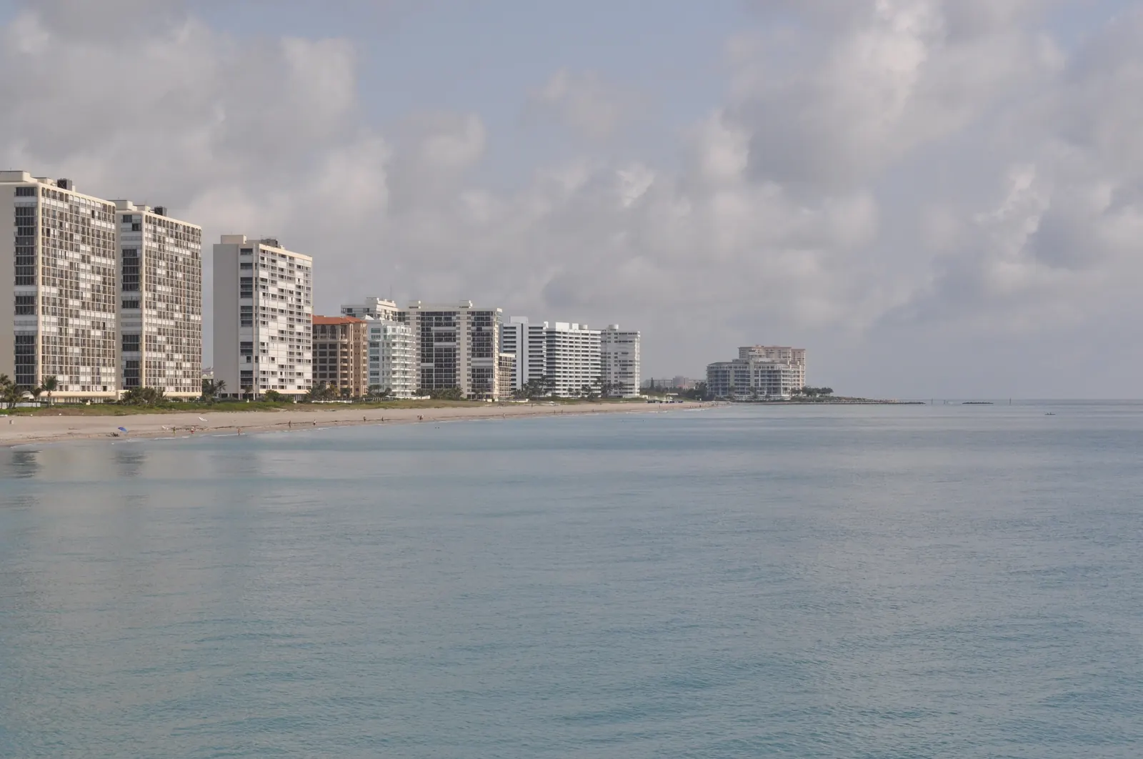 Calm water and a line of high-rise towers along the shore to the north of the Deerfield Beach pier under a cloudy sky.