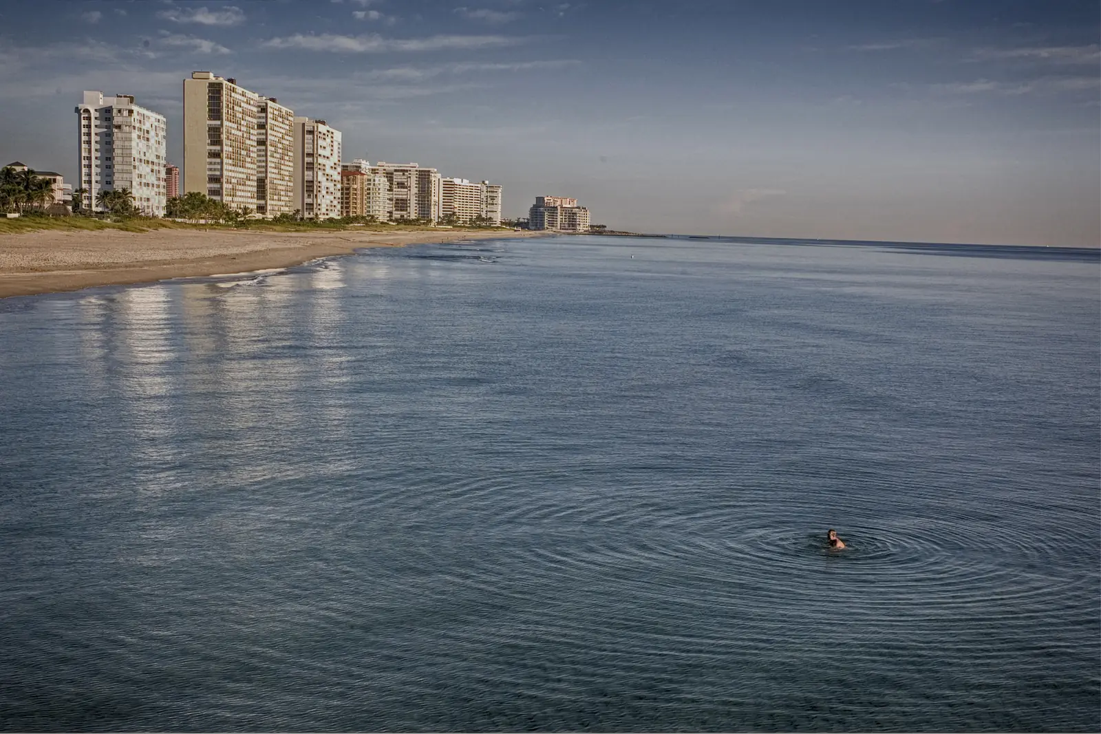 A lone swimmer in still ocean water off Deerfield Beach, with the beach and a row of condominium towers in the distance.