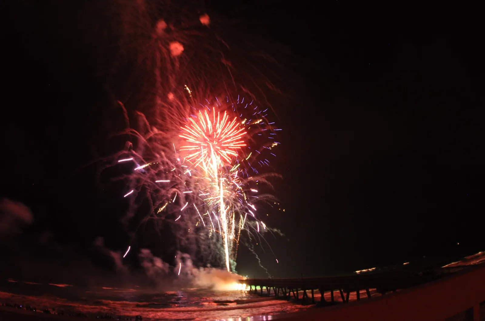 Red and white fireworks burst over the dark ocean beside the Deerfield Beach pier at night.