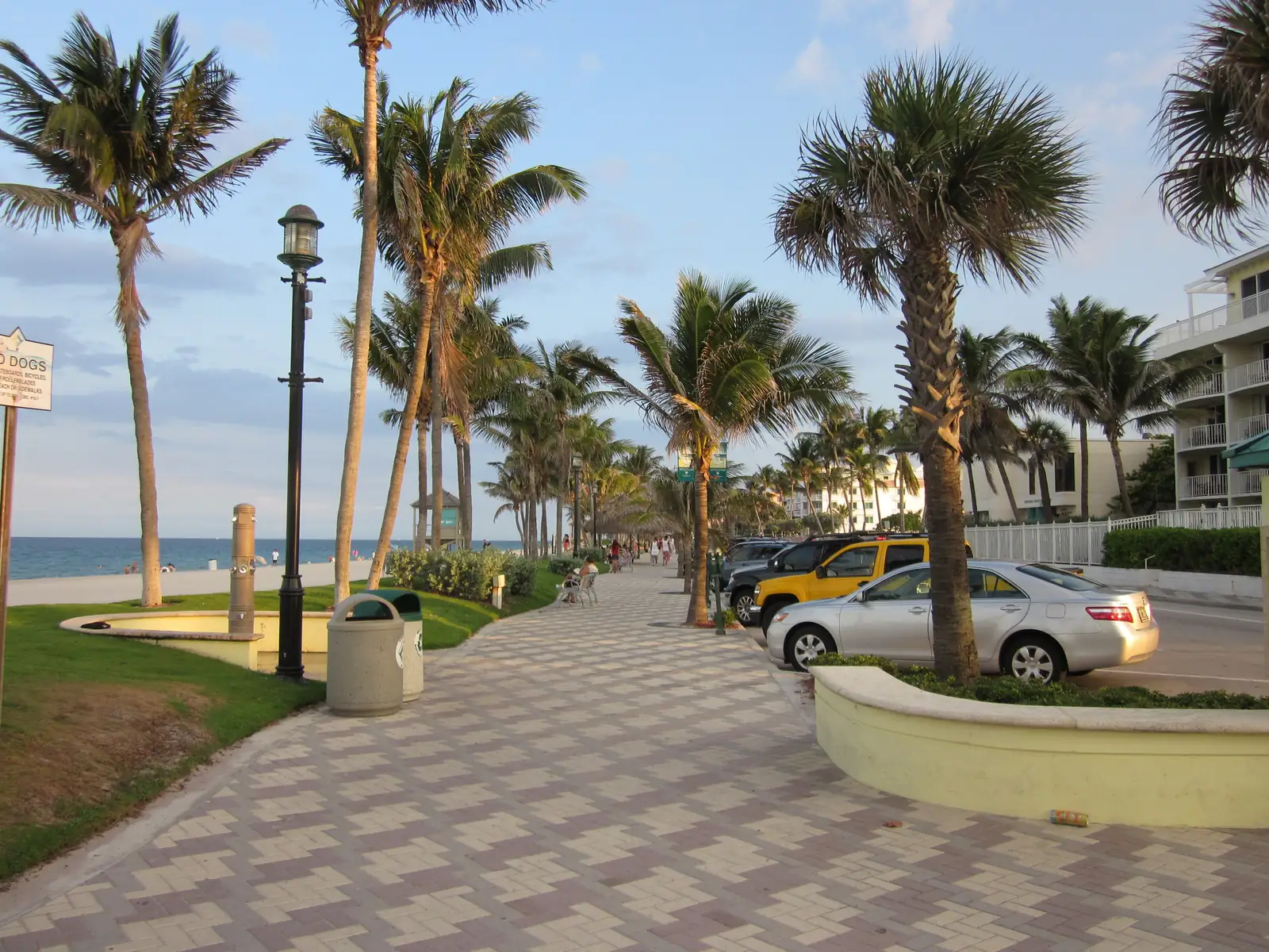 The patterned paver promenade along Deerfield Beach, lined with palms and lamp posts, with parked cars beside it and the beach on the left.