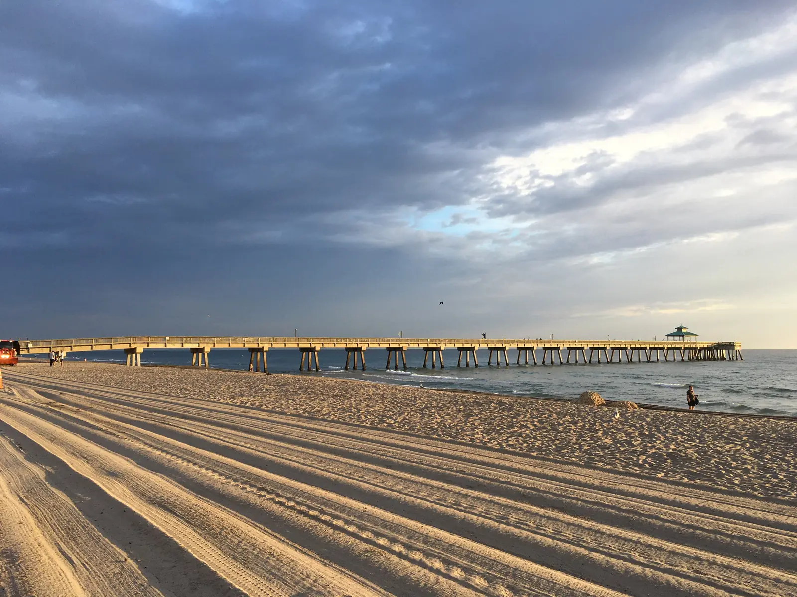 The Deerfield Beach pier running out to sea from a wide, raked beach under a dark cloud bank, with low sun on the sand.