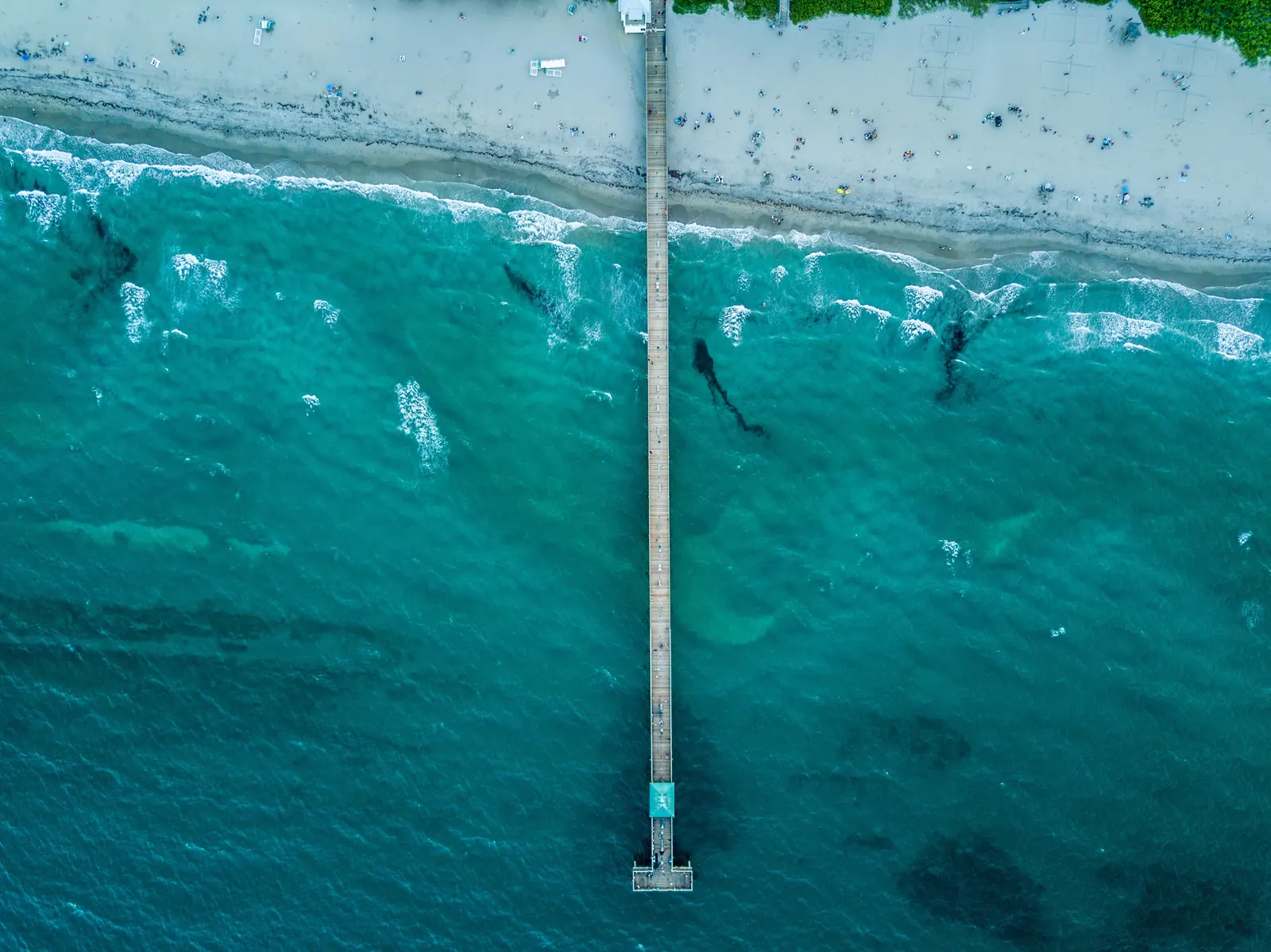Looking straight down on the Deerfield Beach pier from the air: the long deck crosses the surf line and runs out over turquoise water and dark reef patches.