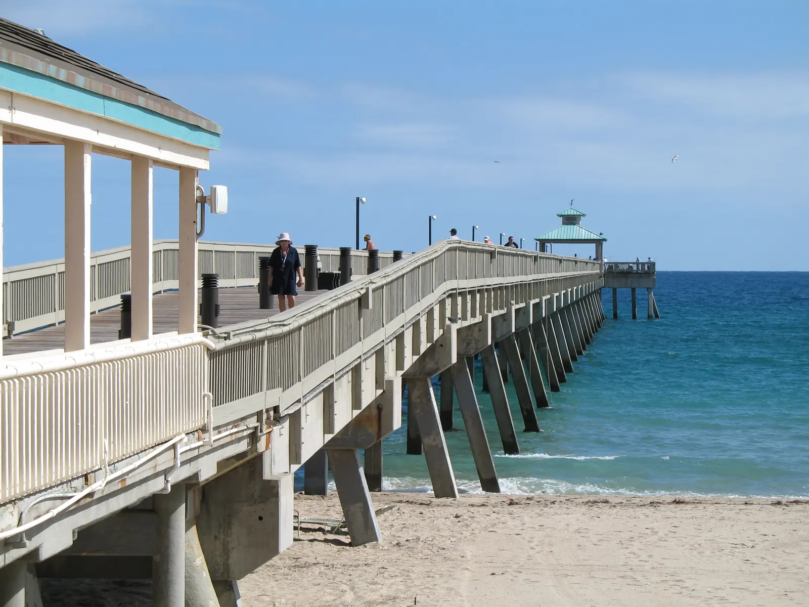 The Deerfield Beach pier rising from the sand on angled concrete supports, with its covered entrance on the left and blue water ahead.