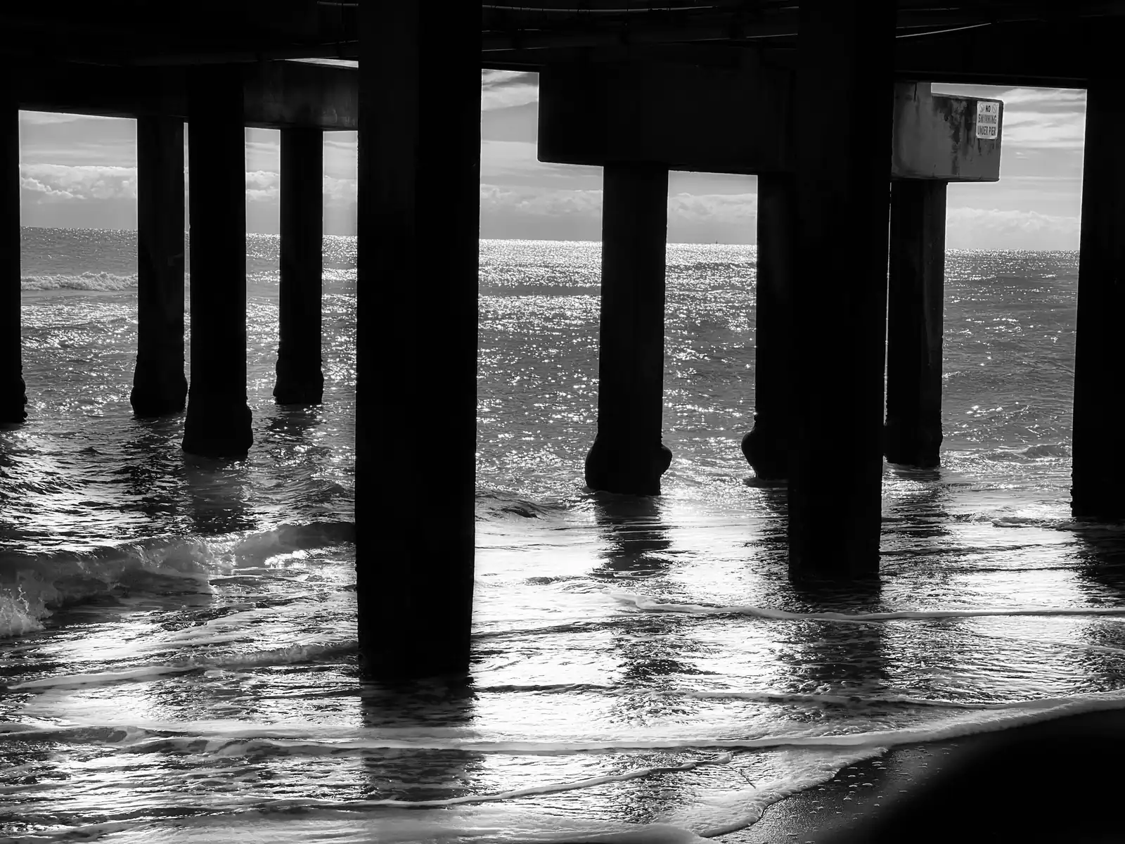 Black and white view from under the Deerfield Beach pier: thick concrete piles stand in the swash with light on the water beyond.