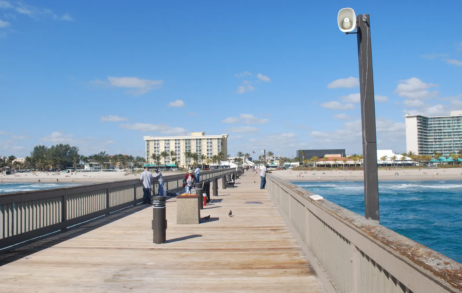 The wooden deck of the Deerfield Beach pier with its railings and a light pole, leading back to the beach and hotels on shore.