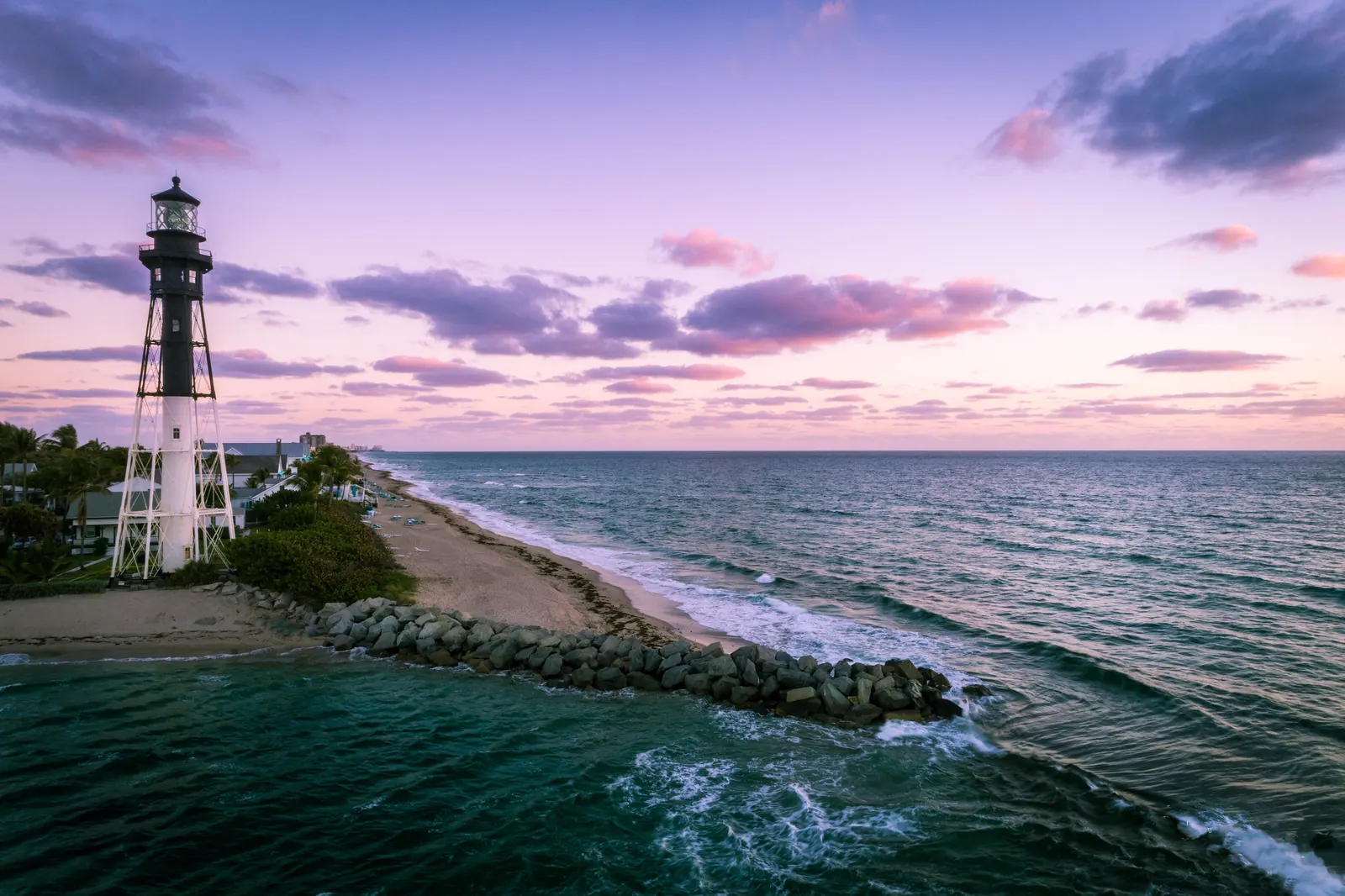 The Hillsboro Inlet Light, a skeletal iron tower painted white below and black above, beside the inlet jetty under a pink dawn sky.