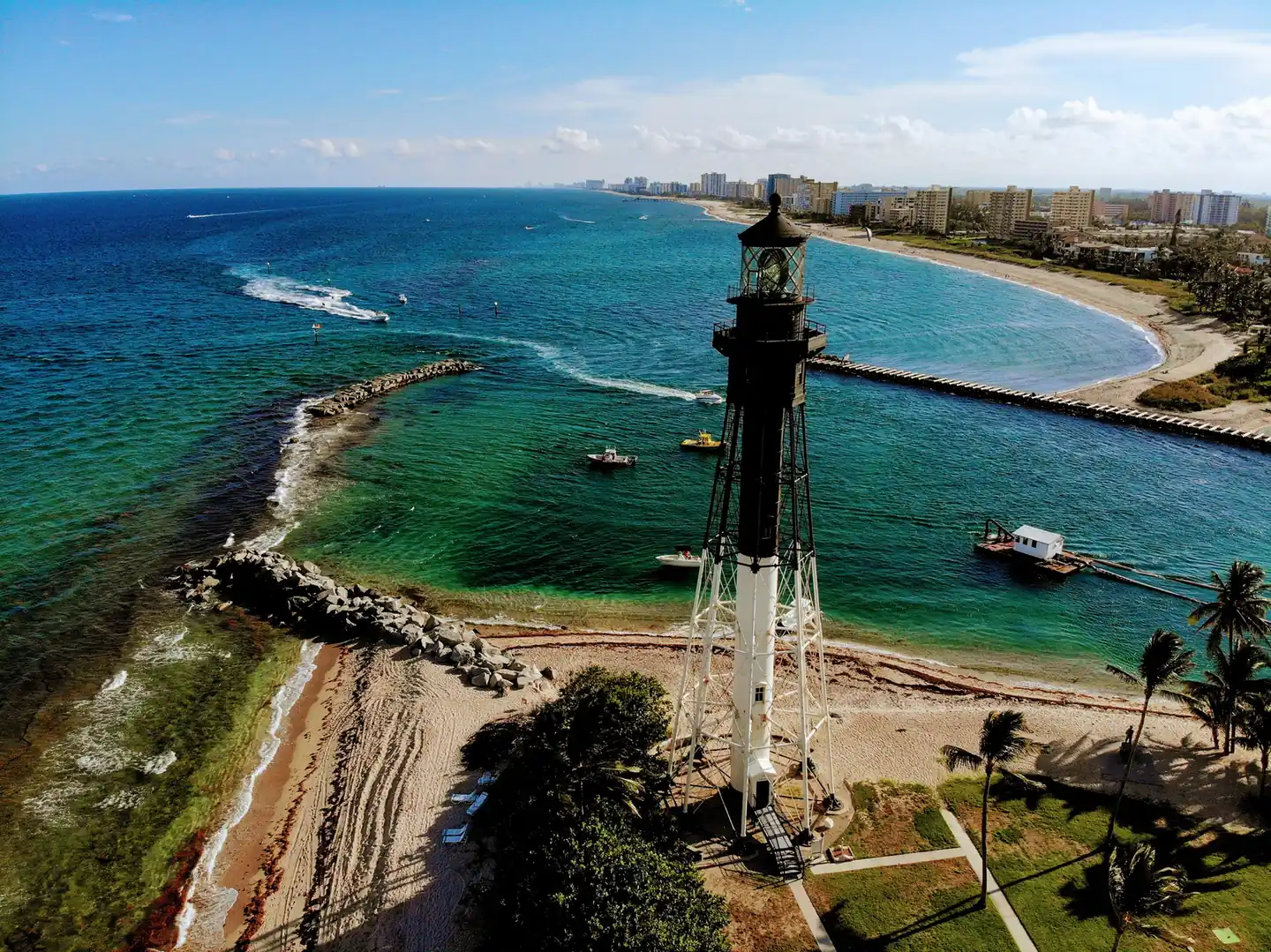 Aerial view of the black and white iron Hillsboro Inlet Light beside the inlet's rock jetty, with boats in green water and the coast curving away.