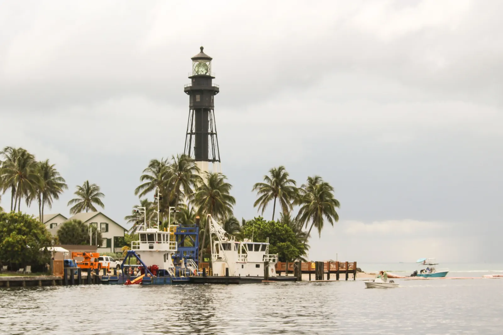 The Hillsboro Inlet Light seen across the water above palm trees, with work boats at a dock in front and a grey sky.