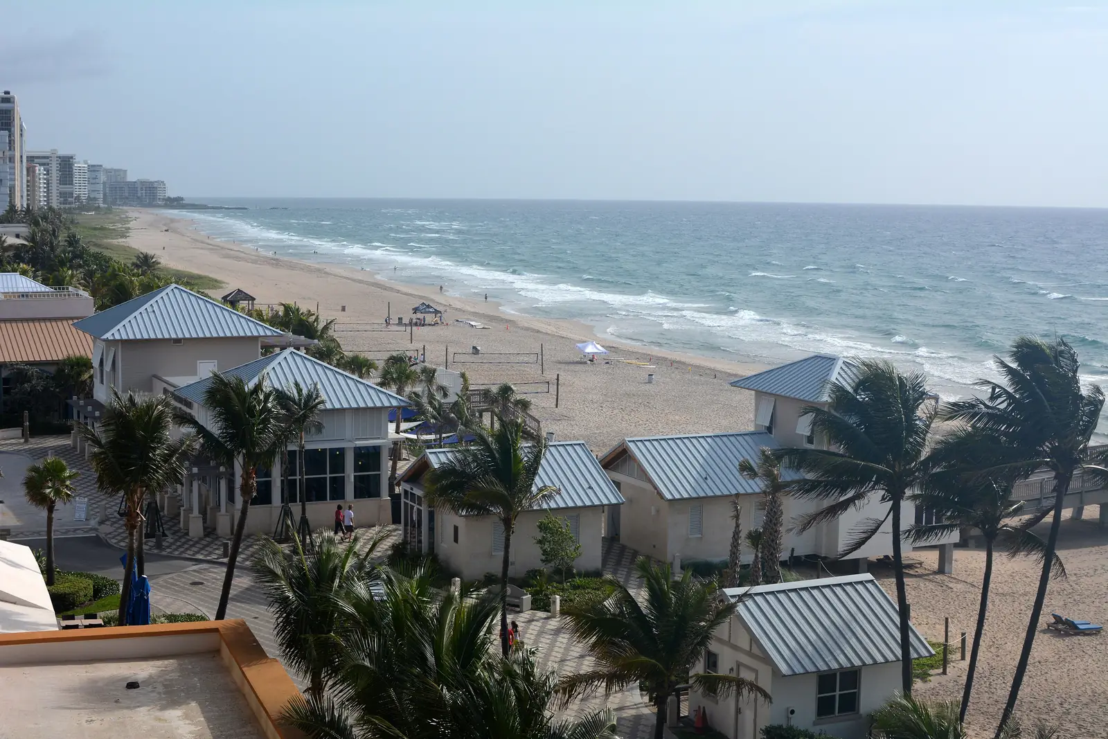 Looking north along Deerfield Beach in early light from a high balcony, over metal-roofed beachfront buildings and palms to the surf.