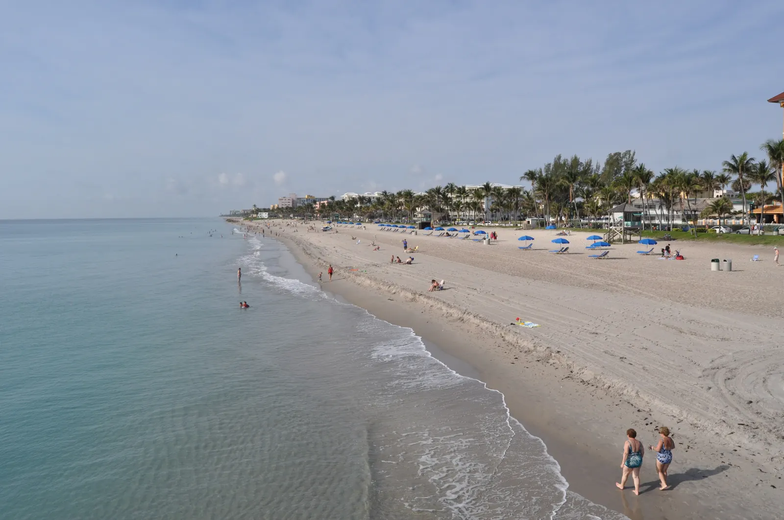 Looking south from the Deerfield Beach pier along the beach, with blue umbrellas, palms and two people walking at the water's edge.